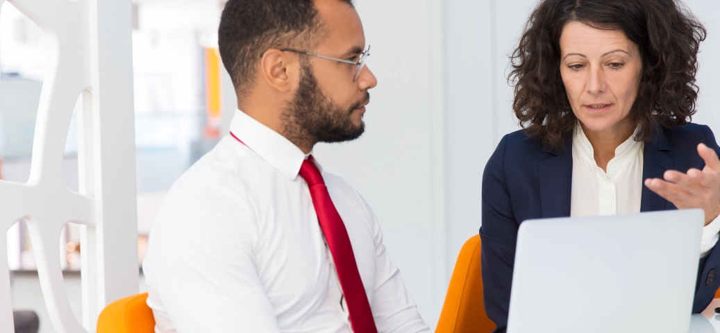 Homem de camisa branca e gravata vermelha escuta atentamente mulher de blazer azul gesticulando em frente ao laptop.