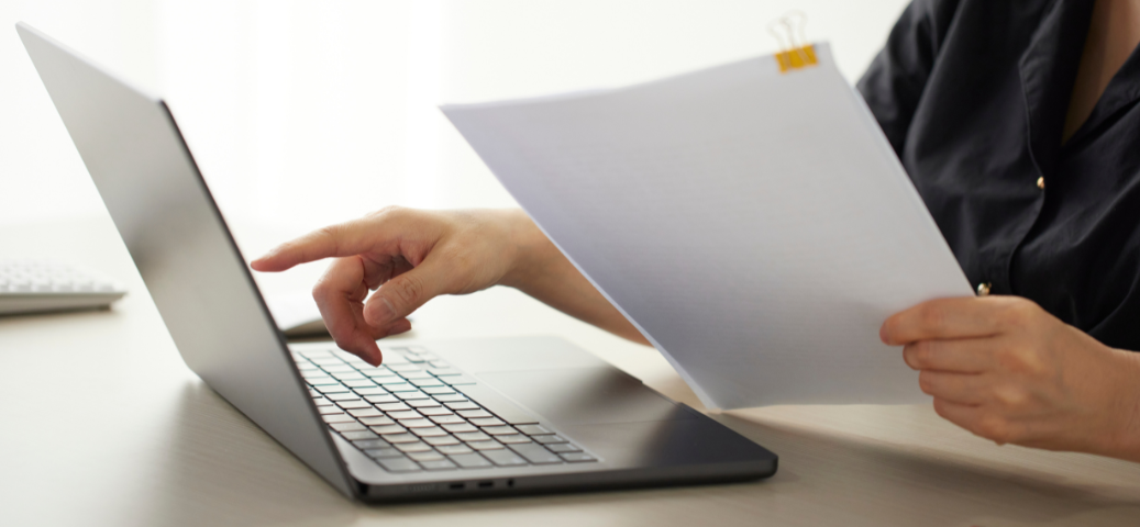 Pessoa aponta para a tela de um laptop enquanto segura papéis presos por um clipe, sugerindo conferência de dados.
