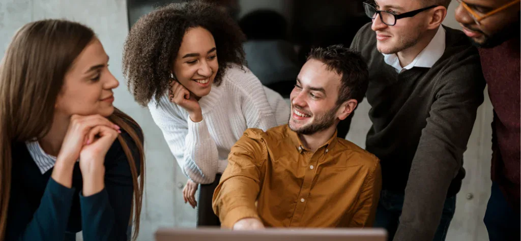 Grupo de quatro colegas de trabalho sorridentes reunidos em volta de um computador em um ambiente de escritório.
