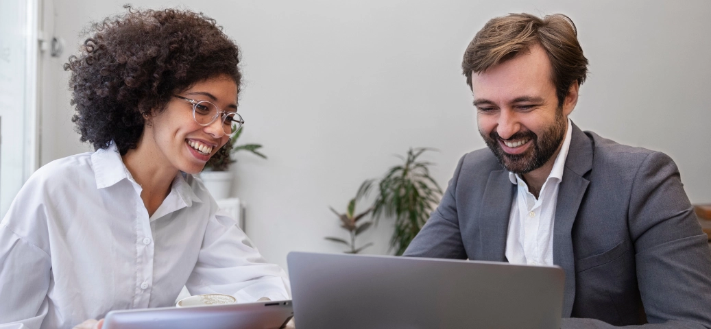 Mulher negra e homem branco sorridentes olham para a tela de um notebook em um ambiente iluminado de escritório.