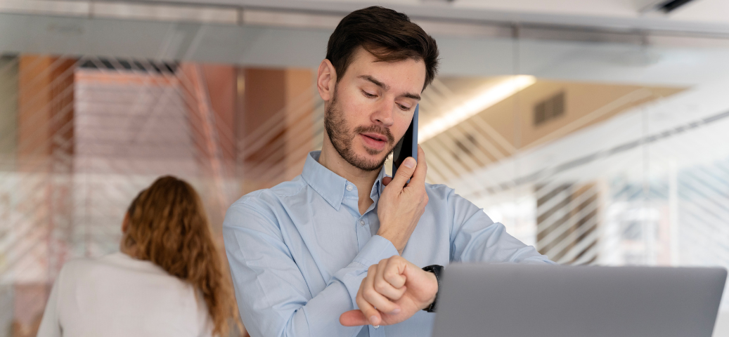 Homem de camisa social azul fala ao celular e olha para o relógio de pulso, demonstrando preocupação com o horário.