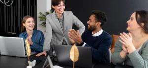 Foto de um ambiente de trabalho positivo. Uma pessoa de blazer cinza está em pé, sorrindo, enquanto três colegas sentados à sua frente a aplaudem com entusiasmo.