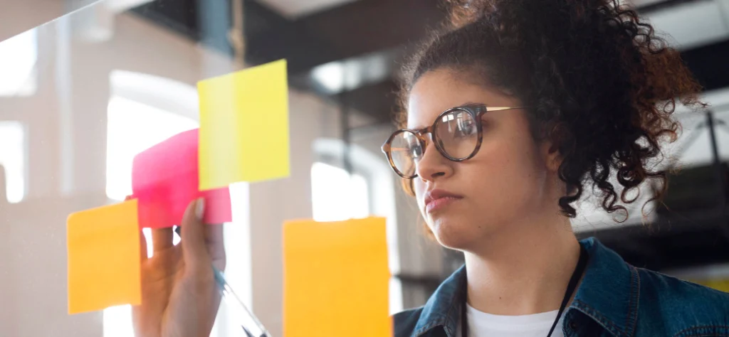 Mulher jovem de óculos observa e organiza notas adesivas coloridas em um painel de vidro durante processo criativo.