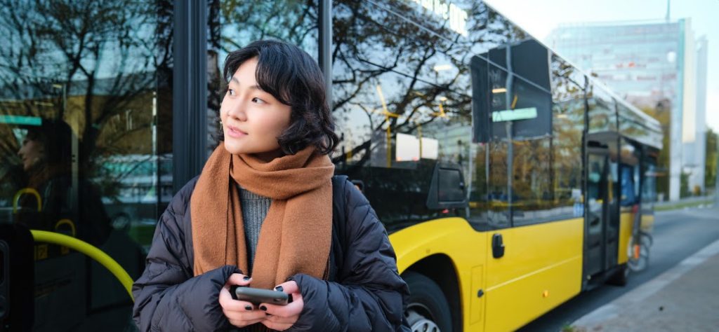 A imagem mostra uma jovem de etnia asiática, com cabelo escuro e curto, em um ambiente urbano. Ela está vestindo um casaco puffer preto sobre um suéter cinza e um cachecol marrom claro bem enrolado no pescoço, o que sugere um clima frio. A jovem está segurando um smartphone com as duas mãos e olhando para cima e para a direita com uma expressão pensativa ou curiosa. Ao fundo, parcialmente visível e ligeiramente desfocado, há um ônibus moderno de cor amarela com detalhes em preto e janelas espelhadas, refletindo árvores e edifícios. A reflexão do ônibus nas janelas parece ter um letreiro digital que, embora ilegível, sugere ser um ônibus de transporte público. O cenário sugere que ela está em uma parada de ônibus ou esperando por um transporte.