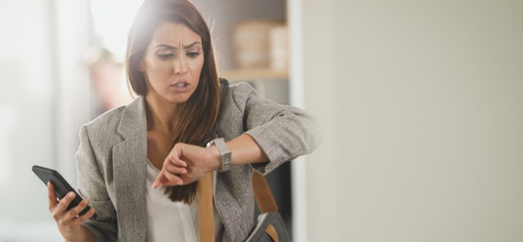 A imagem mostra uma mulher de meia-idade, com cabelos castanhos longos e lisos, vestindo um blazer cinza claro e uma blusa branca. Ela tem uma expressão de preocupação ou surpresa em seu rosto, com as sobrancelhas franzidas e a boca ligeiramente aberta, como se estivesse resmungando ou chocada. Seu olhar está fixo em seu pulso esquerdo, onde ela está olhando um relógio de pulso prateado. Na mão direita, ela segura um smartphone preto. No ombro esquerdo, ela parece carregar uma bolsa escura ou mochila. O fundo está ligeiramente desfocado, mas parece ser um ambiente interno, possivelmente um escritório ou uma casa, com luz natural vindo da esquerda, criando um certo brilho na imagem. A composição sugere que ela está com pressa, atrasada ou surpresa com o horário que vê no relógio.