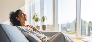 A imagem mostra uma mulher deitada confortavelmente em um sofá cinza, com os olhos fechados e um sorriso relaxado no rosto, como se estivesse desfrutando de um momento de paz ou meditação. Ela tem cabelos escuros e cacheados presos em um rabo de cavalo e veste roupas casuais de cor clara, possivelmente um moletom ou cardigan cinza sobre uma blusa mais clara. A cena se passa em um ambiente interno bem iluminado pela luz natural que entra por uma grande janela ao fundo. Através da janela, é possível ver o exterior, com folhagens verdes e possivelmente outras construções ou árvores. Há duas plantas em vasos de cerâmica claras perto da janela, adicionando um toque de natureza ao ambiente. Um copo com água está visível sobre uma superfície à direita do sofá. A atmosfera geral da imagem transmite tranquilidade e bem-estar.