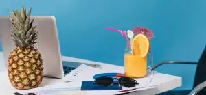 Mesa de escritório com abacaxi, laptop, documentos, óculos de sol e bebida tropical com laranja, canudo e guarda-chuva.
