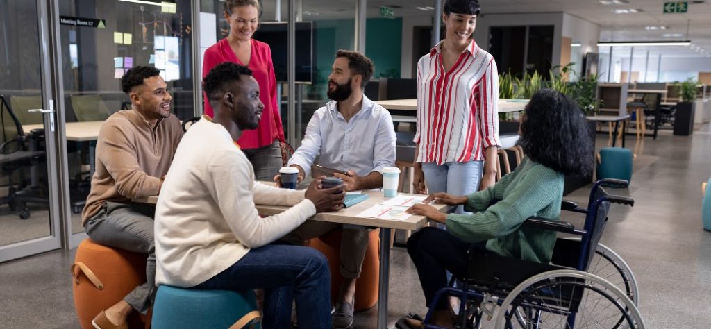 A imagem mostra um grupo diverso de seis adultos em um ambiente de escritório moderno e aberto. Eles estão reunidos em torno de uma pequena mesa de madeira clara, interagindo e parecendo engajados em uma discussão ou reunião informal. Da esquerda para a direita, temos: Um homem de pele escura com um moletom cor de areia e calças cinzas, sentado em um pufe laranja. Ele está sorrindo e olhando para o centro do grupo. Um homem negro de pele escura com uma camisa creme de manga comprida e calça jeans, sentado em um pufe azul-esverdeado. Ele está segurando uma caneca e olhando para o homem à sua frente. Uma mulher branca com uma blusa rosa vibrante e calça escura, que está de pé atrás da mesa e sorrindo para o grupo. Um homem barbudo com pele clara e uma camisa social branca de manga comprida, sentado na mesa e segurando uma caneca. Ele está olhando para a mulher de camisa rosa. Uma mulher com pele clara e cabelo escuro, usando uma camisa listrada vermelha e branca e calça jeans. Ela está de pé do outro lado da mesa, olhando para o centro do grupo. Uma mulher negra sentada em uma cadeira de rodas, usando um suéter verde escuro e calças escuras. Ela está virada para a mesa e parece estar participando ativamente da conversa.