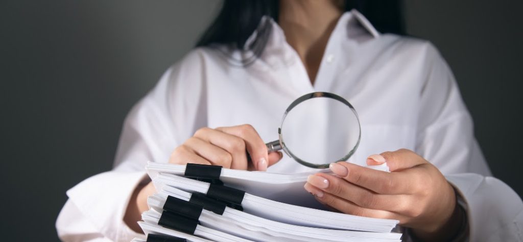 A imagem mostra uma pessoa segurando uma lupa sobre uma pilha de documentos. A pessoa está vestindo uma camisa branca e tem unhas bem cuidadas. A pilha de documentos é grossa e está presa com vários clipes pretos, indicando que são muitos papéis. A lupa está posicionada no centro da imagem, com as mãos da pessoa segurando-a logo abaixo. O fundo é escuro e desfocado, o que faz com que o foco principal da imagem esteja na lupa e nos documentos. A iluminação é suave, destacando os objetos em primeiro plano. A imagem sugere um tema de pesquisa, análise, auditoria ou verificação de informações.