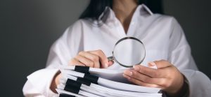 A imagem mostra uma pessoa segurando uma lupa sobre uma pilha de documentos. A pessoa está vestindo uma camisa branca e tem unhas bem cuidadas. A pilha de documentos é grossa e está presa com vários clipes pretos, indicando que são muitos papéis. A lupa está posicionada no centro da imagem, com as mãos da pessoa segurando-a logo abaixo. O fundo é escuro e desfocado, o que faz com que o foco principal da imagem esteja na lupa e nos documentos. A iluminação é suave, destacando os objetos em primeiro plano. A imagem sugere um tema de pesquisa, análise, auditoria ou verificação de informações.