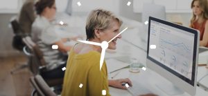A imagem mostra um ambiente de escritório com uma mulher de meia-idade, com cabelos loiros curtos, sorrindo enquanto olha para um monitor de computador à sua frente. Ela veste uma blusa amarela. À esquerda da mulher, há outras pessoas desfocadas trabalhando em suas mesas, e à direita, outra pessoa desfocada pode ser vista ao fundo. A tela do computador exibe gráficos e dados, provavelmente relacionados a finanças ou relatórios. Sobreposta à imagem, há uma representação de um relógio analógico branco, com ponteiros que indicam aproximadamente 10 horas e 10 minutos (o ponteiro das horas está um pouco antes do 2 e o dos minutos está no 2).