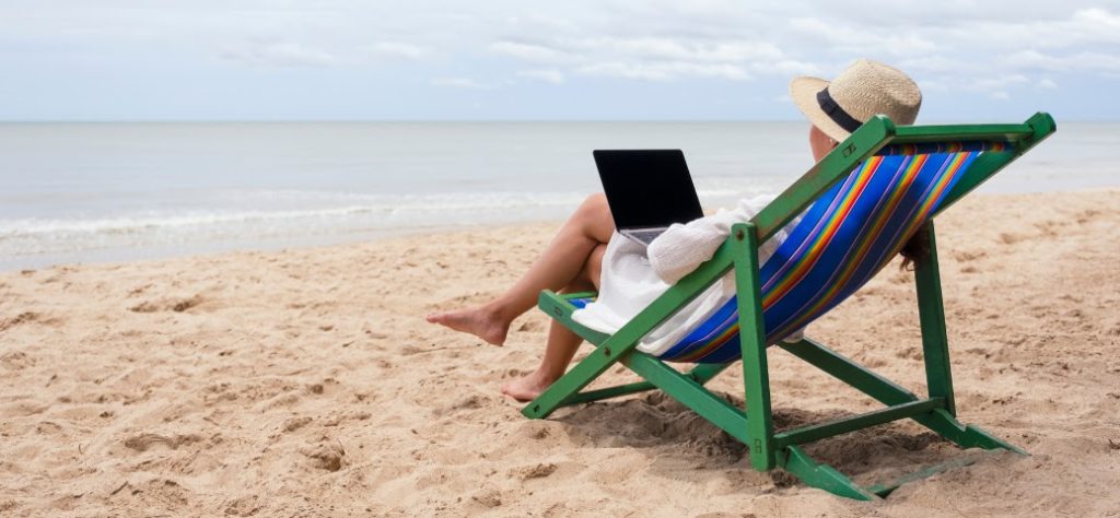 A imagem mostra uma pessoa sentada em uma cadeira de praia listrada, de costas para o observador, com vista para o oceano. A pessoa está usando um chapéu de palha e uma roupa clara, e parece estar utilizando um laptop que está apoiado em seu colo. A cena se passa em uma praia, com areia clara em primeiro plano e as ondas do mar suaves ao fundo. O céu está parcialmente nublado, e a linha do horizonte entre o mar e o céu é visível. A cadeira tem uma estrutura de madeira verde e um tecido com listras coloridas em azul, branco, amarelo, laranja e verde. A impressão geral é de relaxamento e trabalho remoto em um ambiente de férias.