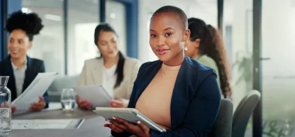 Uma mulher com um blazer claro opera uma calculadora enquanto trabalha em um laptop, com documentos financeiros em uma mesa de madeira em um escritório iluminado.
