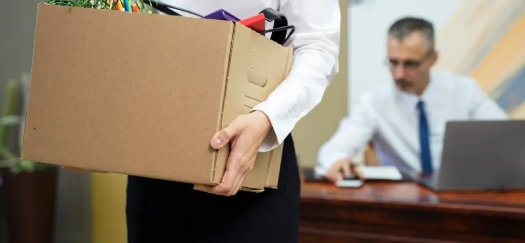 Mulher demitida segura uma caixa de papelão com seus pertences de escritório, enquanto um homem de terno, possivelmente o chefe, está sentado à mesa atrás dela.