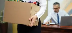 Mulher demitida segura uma caixa de papelão com seus pertences de escritório, enquanto um homem de terno, possivelmente o chefe, está sentado à mesa atrás dela.
