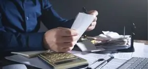 Mãos de uma pessoa segurando um pequeno pedaço de papel branco, possivelmente um recibo. Ao fundo, uma mesa bagunçada com uma pilha de recibos e uma calculadora dourada.