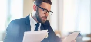 Um homem barbudo de terno e óculos lendo papéis em um escritório.