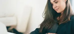 Uma mulher grávida com cabelo castanho escuro e uma blusa cinza, vestindo um blazer preto, está sentada em uma mesa e olha para baixo com uma expressão calma e gentil. Ela apoia a mão na barriga e parece estar tocando na tela de um notebook que não aparece na imagem.