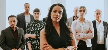 Mulher negra confiante no centro da imagem, com os braços cruzados, liderando um grupo diversificado de profissionais em segundo plano.