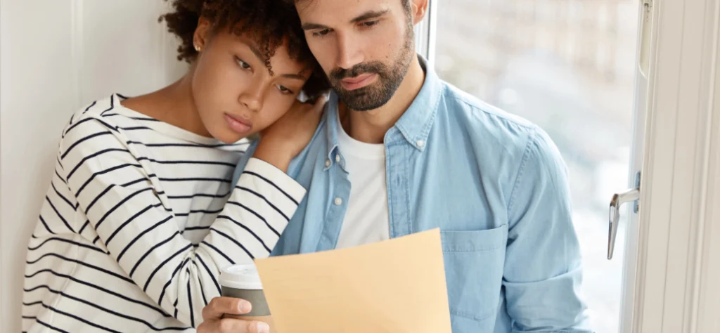 Um casal está em um momento de reflexão, lendo um documento. A mulher, de cabelos curtos, apoia a cabeça no ombro do homem e segura um copo de café. O homem, com barba, segura um envelope e olha para o documento. Ambos parecem concentrados e com semblante sério. A cena se passa em um ambiente com uma janela grande ao fundo.