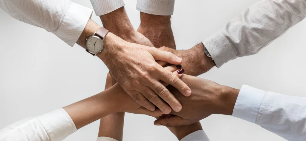 As mãos de cinco pessoas se unem, uma sobre a outra, em um gesto de união e trabalho em equipe. As pessoas vestem camisas de manga comprida, predominantemente brancas. Uma delas usa um relógio de pulso. As unhas de uma das pessoas estão pintadas com esmalte escuro.