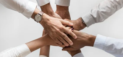 As mãos de cinco pessoas se unem, uma sobre a outra, em um gesto de união e trabalho em equipe. As pessoas vestem camisas de manga comprida, predominantemente brancas. Uma delas usa um relógio de pulso. As unhas de uma das pessoas estão pintadas com esmalte escuro.