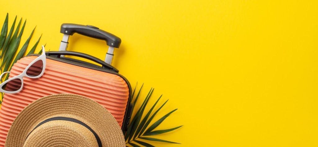 Uma mala de viagem cor de salmão, óculos de sol, um chapéu de palha e folhas de palmeira em um fundo amarelo vibrante.