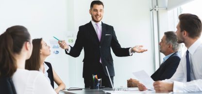 Um grupo de profissionais sentados em uma mesa de reunião, ouvindo um colega em pé que gesticula com as mãos.