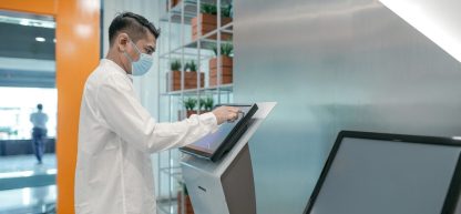 Um homem com máscara médica e um jaleco interage com um quiosque de autoatendimento de ponto, digitando na tela sensível ao toque.