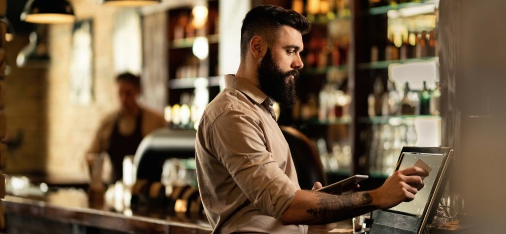 Um bartender barbudo e concentrado toca a tela de um caixa eletrônico em um bar movimentado.
