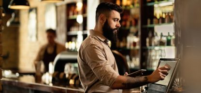 Um bartender barbudo e concentrado toca a tela de um caixa eletrônico em um bar movimentado.