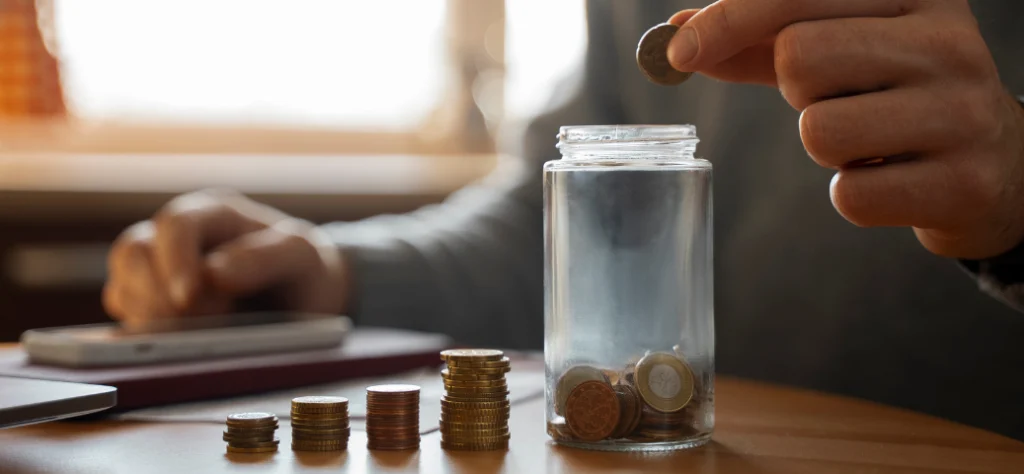 Close-up de uma mão colocando uma moeda em um pote de vidro com moedas, ao lado de pilhas de moedas crescentes.