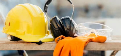 Equipamentos de Proteção Individual (EPIs) dispostos sobre uma superfície de madeira, incluindo um capacete de segurança amarelo, abafadores de ruído pretos, óculos de proteção transparentes e uma luva de trabalho laranja.