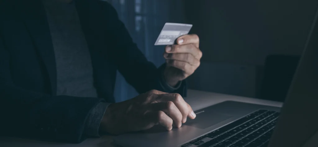 Homem em ambiente escuro, segurando um cartão de crédito e digitando em um notebook, sugerindo fraude ou hacking.