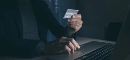 Homem em ambiente escuro, segurando um cartão de crédito e digitando em um notebook, sugerindo fraude ou hacking.