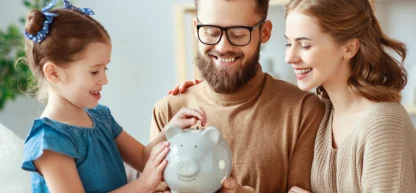 Família ensinando educação financeira: pai, mãe e filha sorrindo enquanto colocam moedas em um cofrinho de porco.