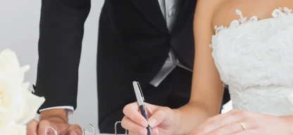 Mão da noiva assinando documentos de casamento (registro civil ou certidão), com o noivo de terno ao lado.