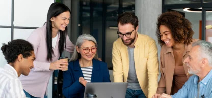 Equipe intergeracional rindo e colaborando em torno de um laptop. Mentoria e troca de experiências no trabalho.