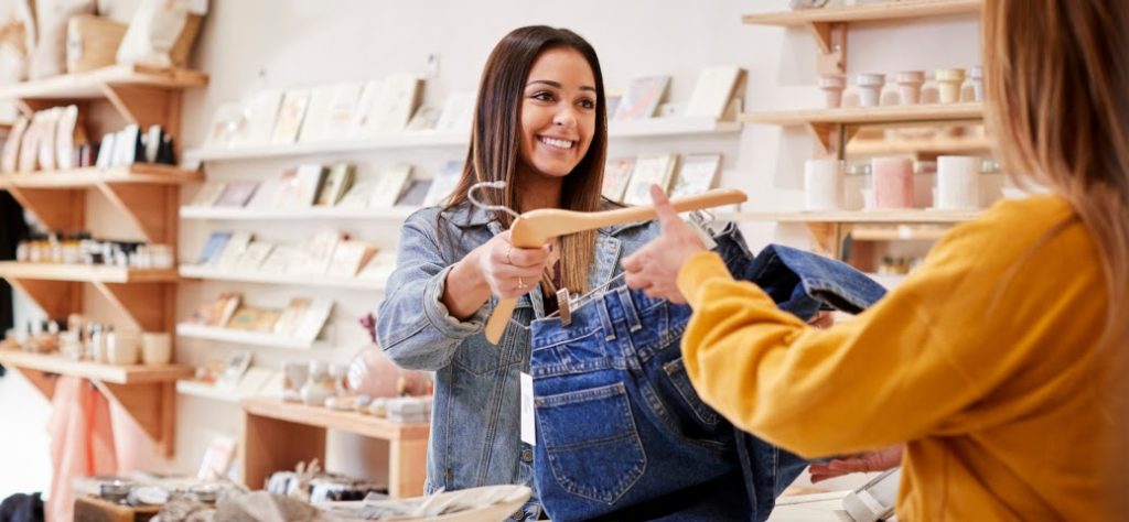 Uma vendedora sorridente em uma loja entrega um par de jeans em um cabide de madeira para uma cliente, simbolizando uma venda.