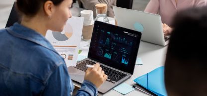 Mulher em jaqueta jeans analisando gráficos e dados de negócios em um notebook em uma reunião.