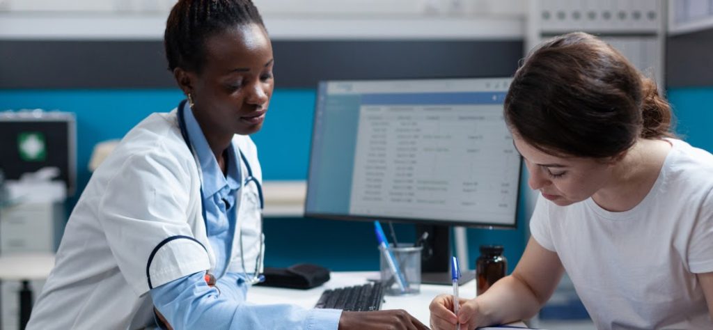 Médica e paciente. Mulher negra de jaleco branco atende mulher branca que preenche formulário em consultório médico.
