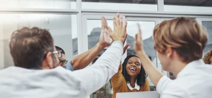 Um grupo diversificado de quatro colegas de trabalho dá um "high five" animado em um escritório moderno, celebrando o sucesso da equipe.