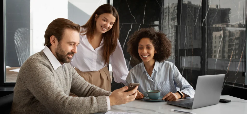 Três colegas de trabalho, dois homens e uma mulher, sorriem enquanto olham para um smartphone na mesa de um escritório moderno.