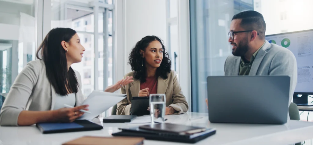 Três executivos (duas mulheres e um homem) em reunião de negócios. Comunicação organizacional e discussão de ideias.