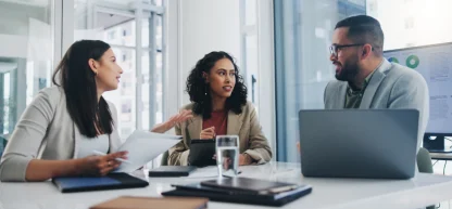 Três executivos (duas mulheres e um homem) em reunião de negócios. Comunicação organizacional e discussão de ideias.