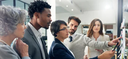 Equipe diversificada de profissionais em reunião, olhando para um quadro com post-its. Estrutura e planejamento.