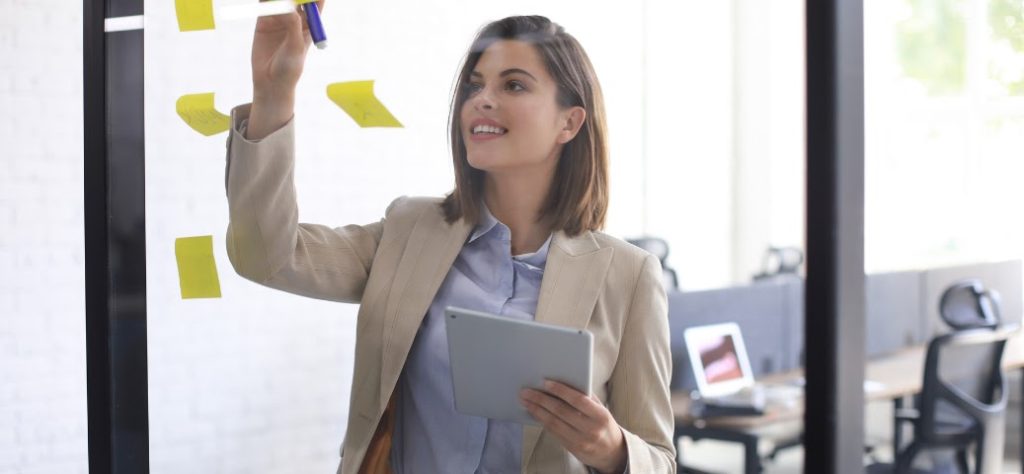 Mulher em traje executivo, com um tablet na mão, anota em post-its amarelos fixados em um painel de vidro no escritório.