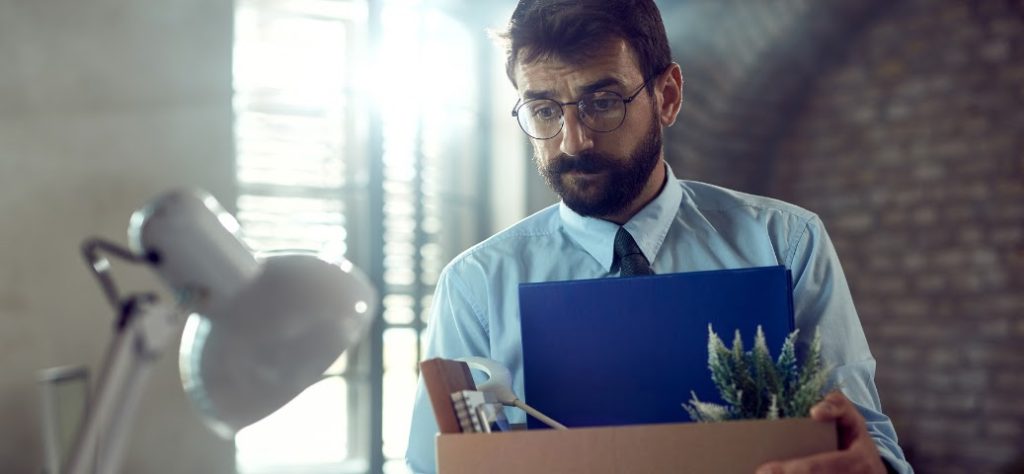 Homem de barba e óculos, com expressão cabisbaixa, segura uma caixa de papelão com pertences (incluindo uma planta) ao deixar o escritório.