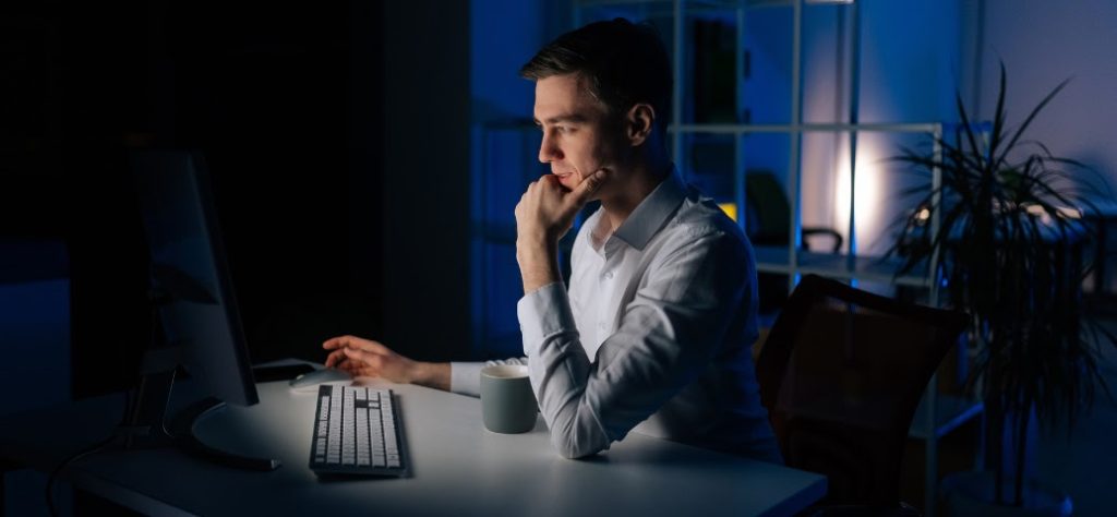 Homem em camisa social branca, trabalhando no computador em um escritório escuro, com iluminação apenas da tela e um abajur discreto.