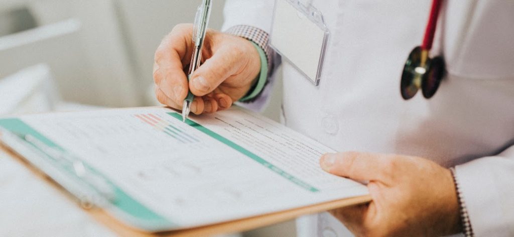 Mãos de um médico (ou profissional de saúde) em jaleco branco preenchendo ou assinando um documento em uma prancheta.